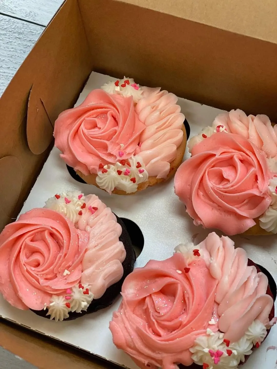 Four heart-shaped cupcakes in a cardboard box, each topped with pink and cream swirled frosting rosettes and small red and pink heart sprinkles.