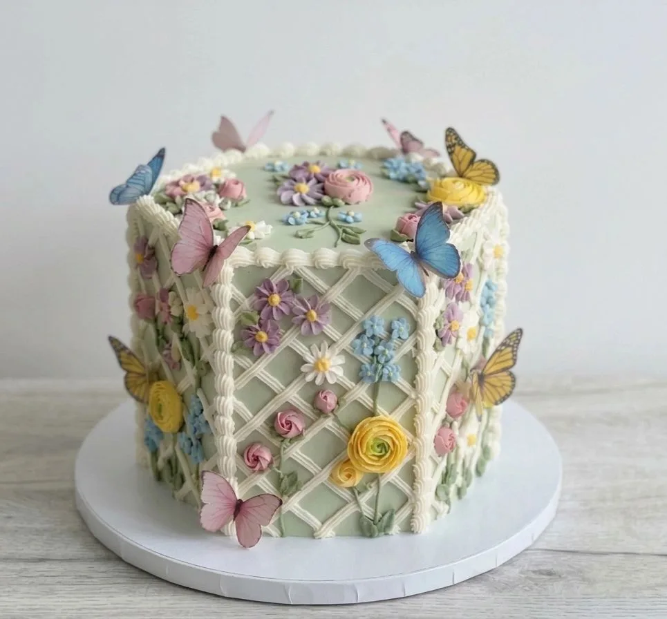 Sage green round cake decorated with buttercream lattice piping, pastel flowers, and pink, blue, and yellow butterflies on a white cake board.