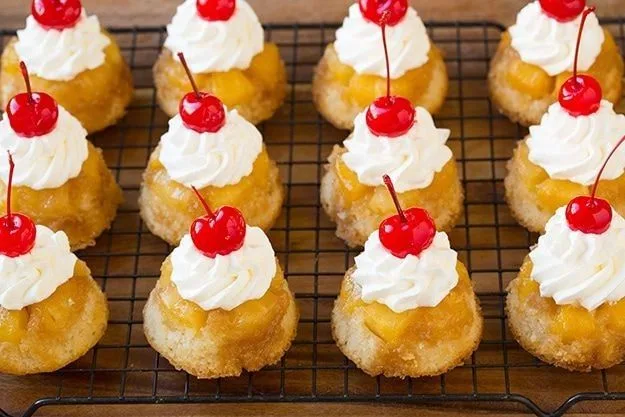Mini desserts with caramelized golden bases, piped whipped cream, and red cherries arranged on a black wire cooling rack.