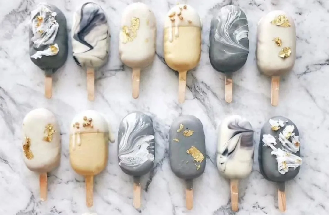 Twelve marbled gray, white, and gold-decorated popsicles with wooden sticks arranged in two rows on marble.