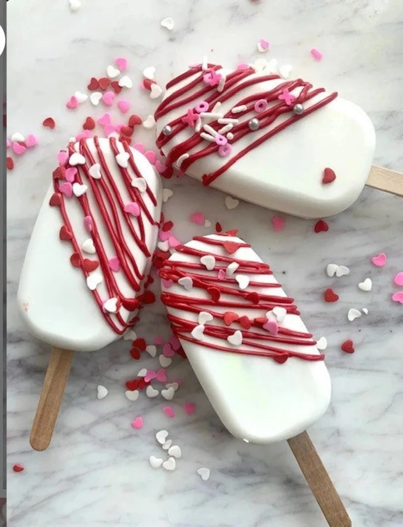 Three white chocolate-covered popsicles on wooden sticks, drizzled with red icing and topped with pink, red.