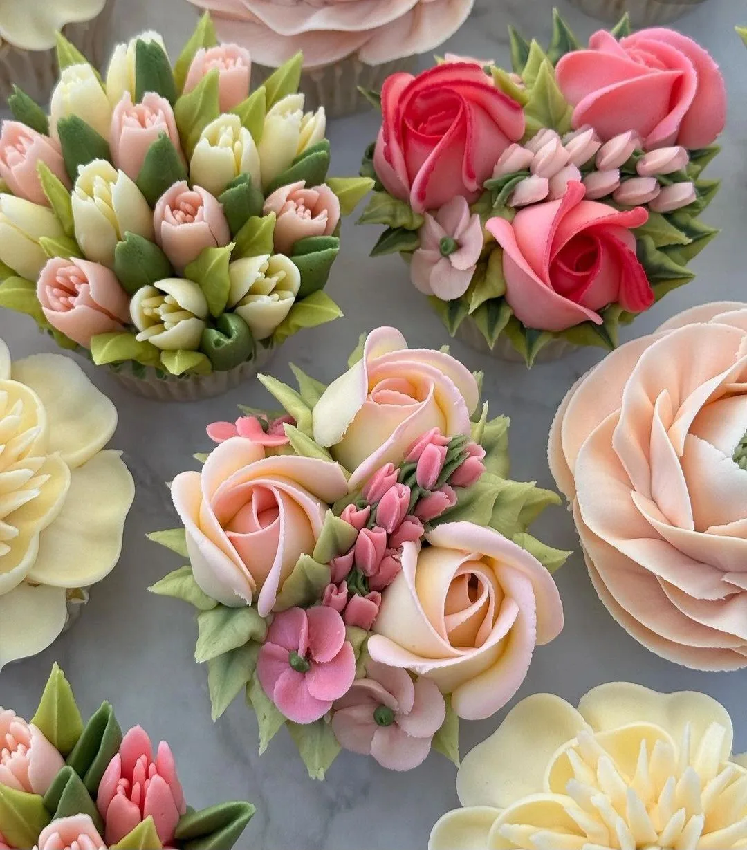 Cupcakes topped with piped buttercream roses, blossoms, and leaves in pink, peach, cream, and green shades.