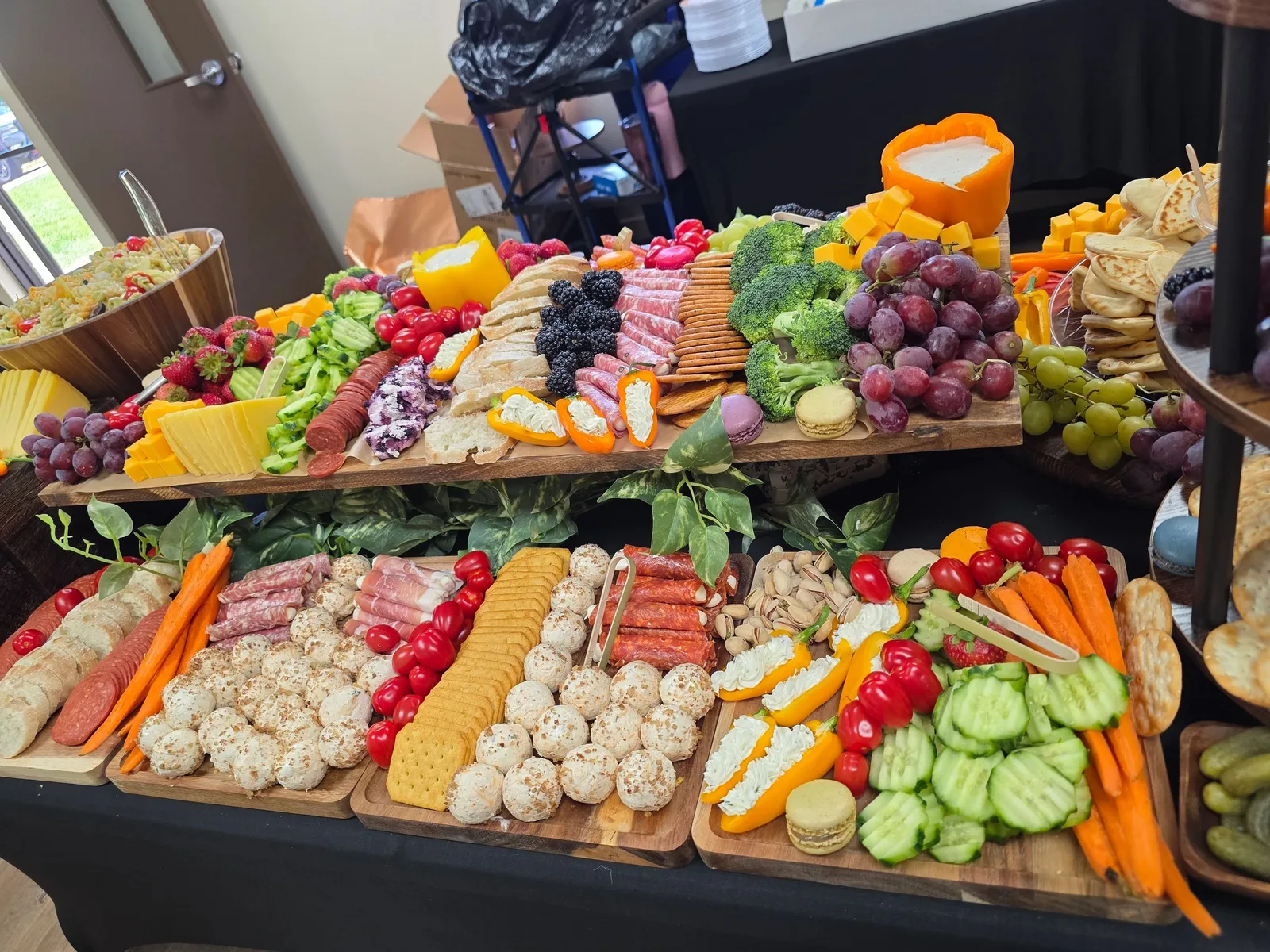 Grazing table on black tablecloth with cheeses, meats, fruits, vegetables, crackers, and macarons on wooden boards.
