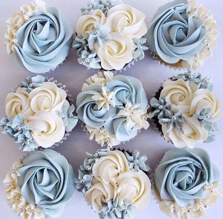 Nine cupcakes decorated with piped buttercream rosettes in dusty blue and cream colors, accented with silver edible pearls.