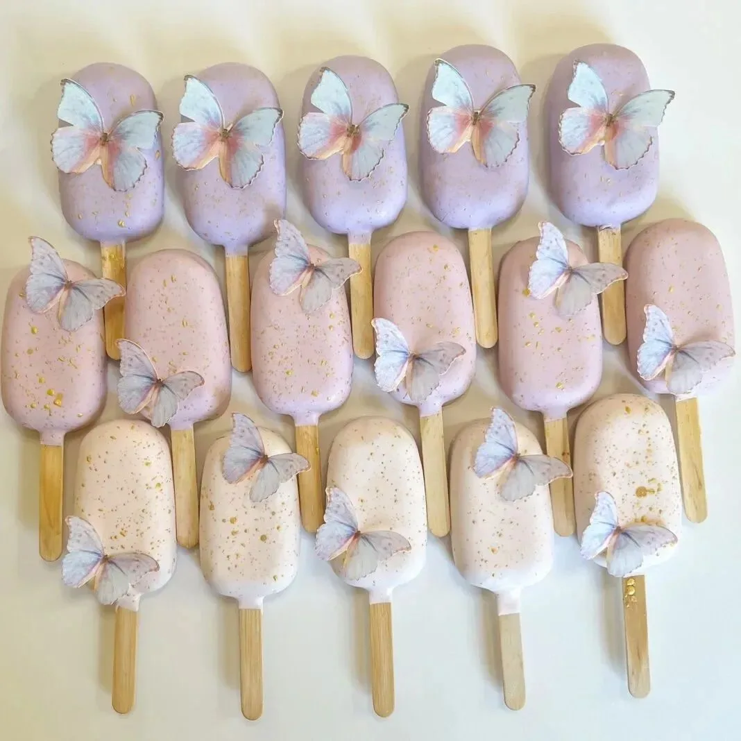 Rows of pastel purple, pink, and cream cake pops decorated with gold speckles and edible butterflies on wooden sticks.