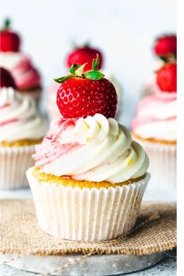 A cupcake with white and pink swirled frosting topped with a fresh whole strawberry, served in a white paper liner.