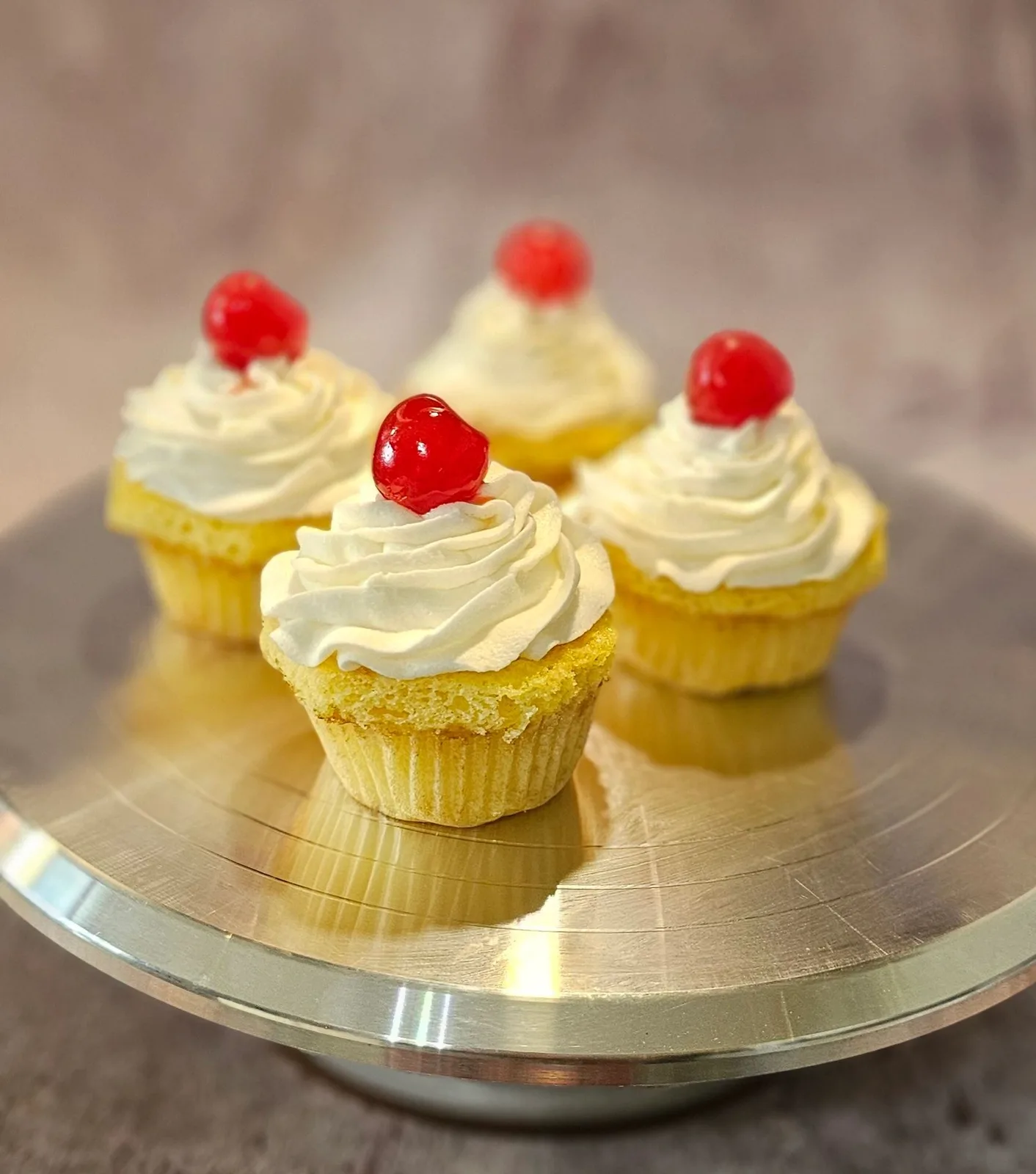 Four yellow cupcakes topped with swirled white frosting and red cherries, displayed on a gold cake stand.