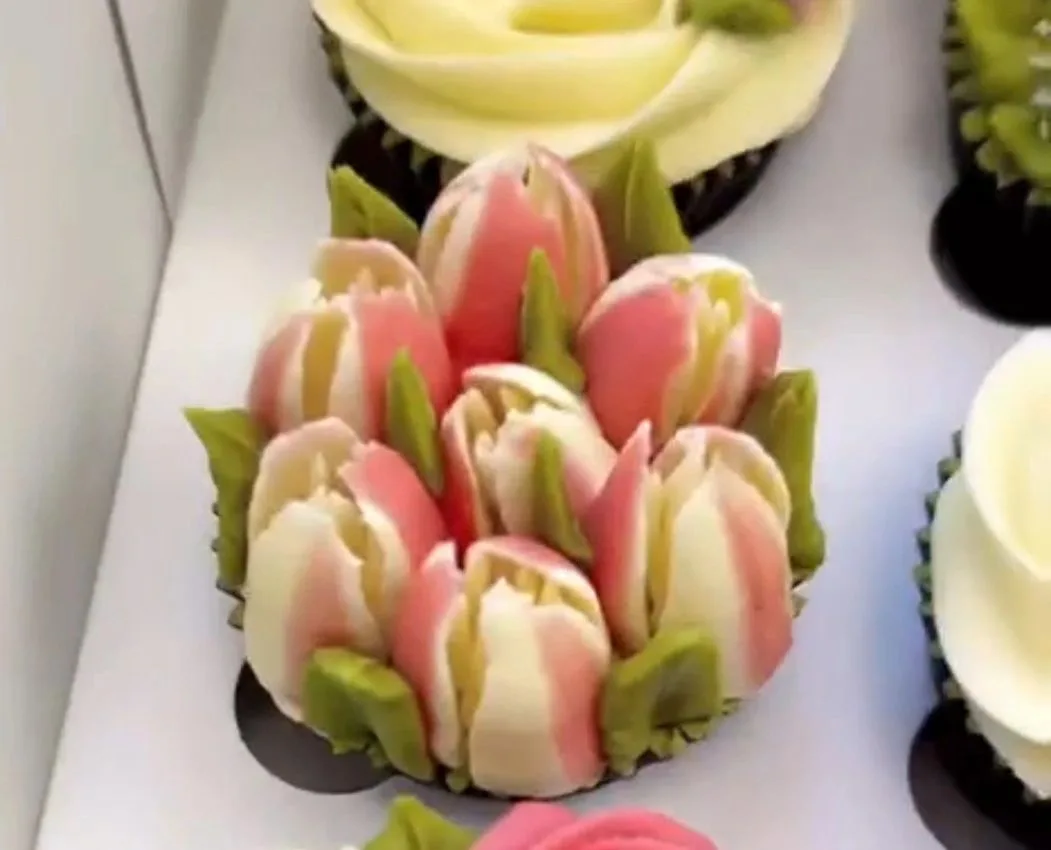 A cupcake decorated with piped white and pink tulip-shaped frosting surrounded by green leaves, alongside other frosted cupcakes.