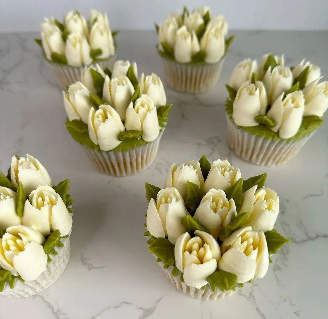 White frosted cupcakes decorated as tulip bouquets with green leaves, displayed on a marble surface.