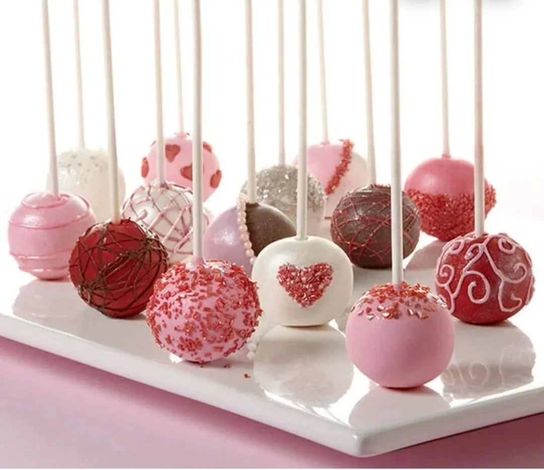 Assorted pink, red, white, and chocolate cake pops decorated with sprinkles, hearts, and drizzle arranged on a white square plate.