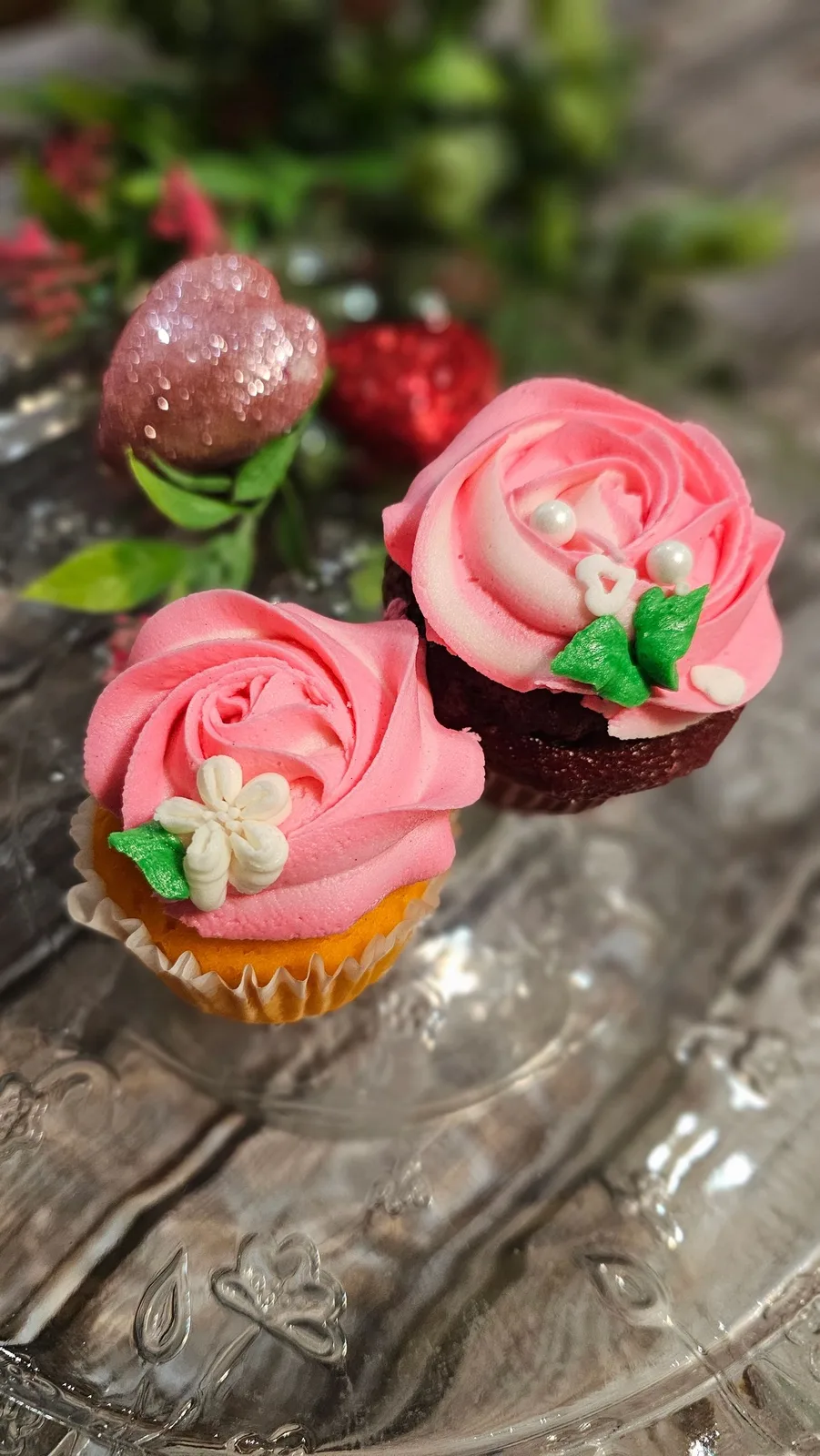 Two cupcakes rest on a clear, patterned glass stand, each topped with a piped pink frosting rose coated in coarse sugar.