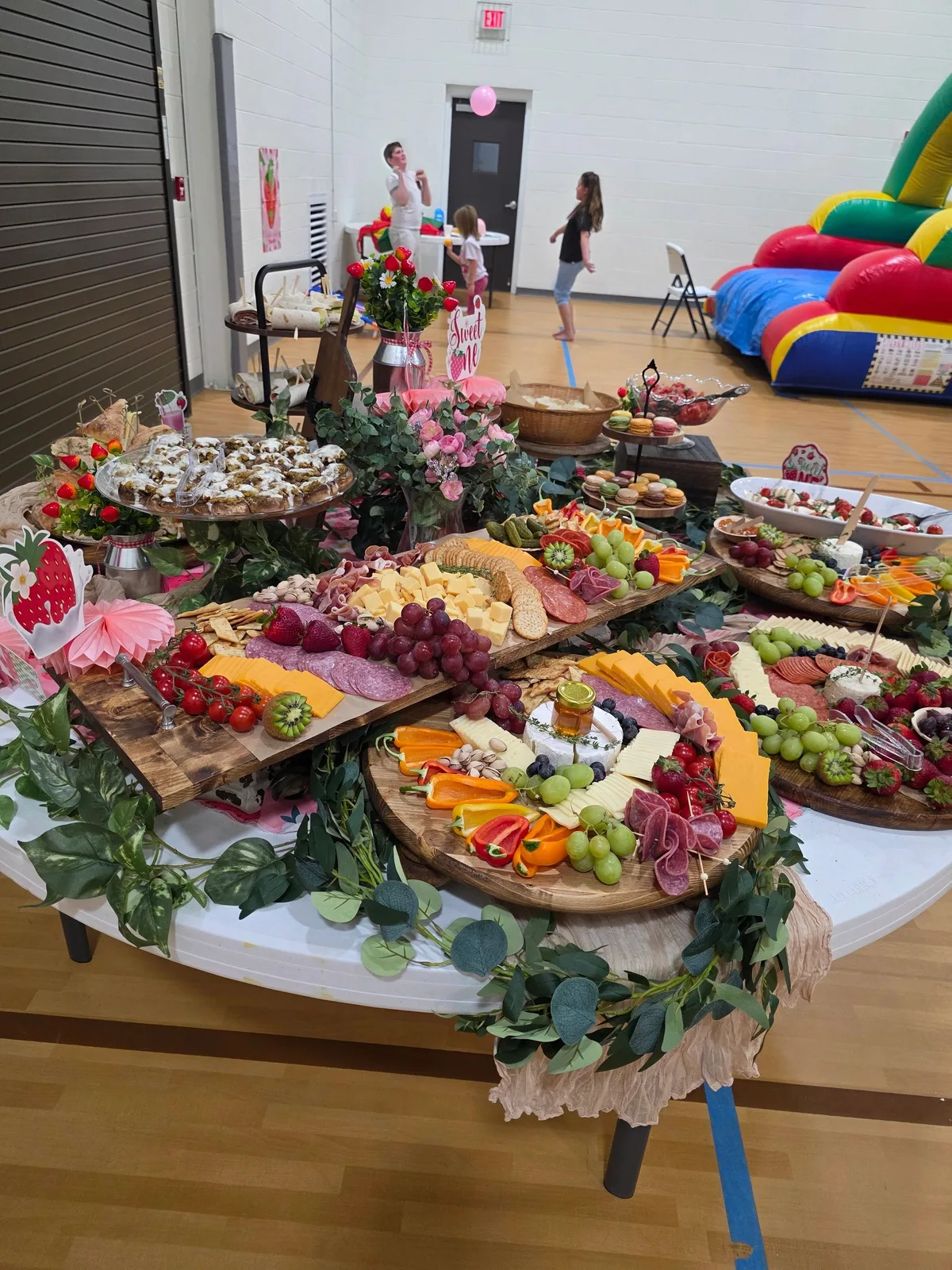 A party grazing table with charcuterie boards, pastries, and floral decor set up in a gymnasium with a bounce house.