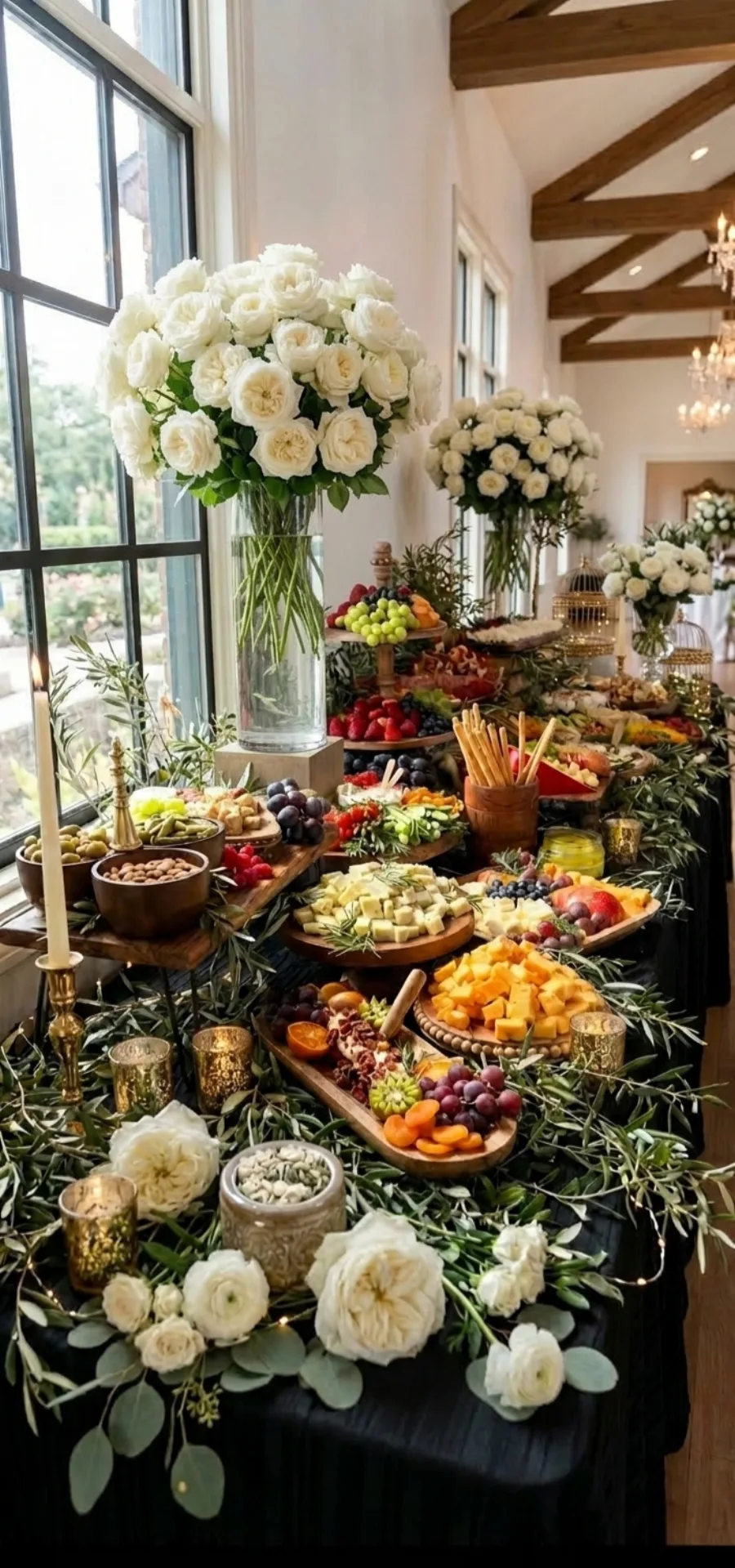 Grazing table set along a window with cheeses, cured meats, fresh and dried fruit, olives, breadsticks, candles and white roses on a black cloth.