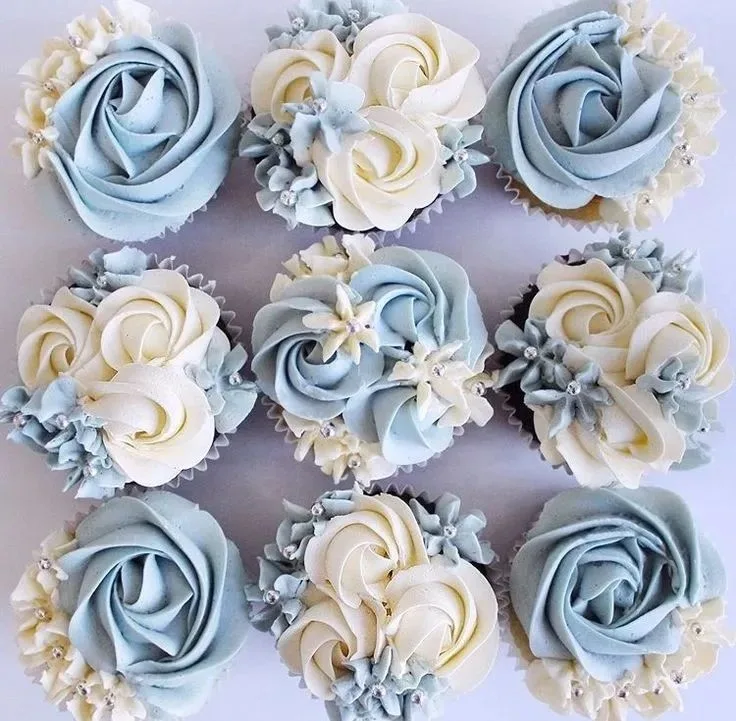 Nine cupcakes decorated with pale blue and white frosting rosettes, small floral accents, and silver pearl sprinkles.