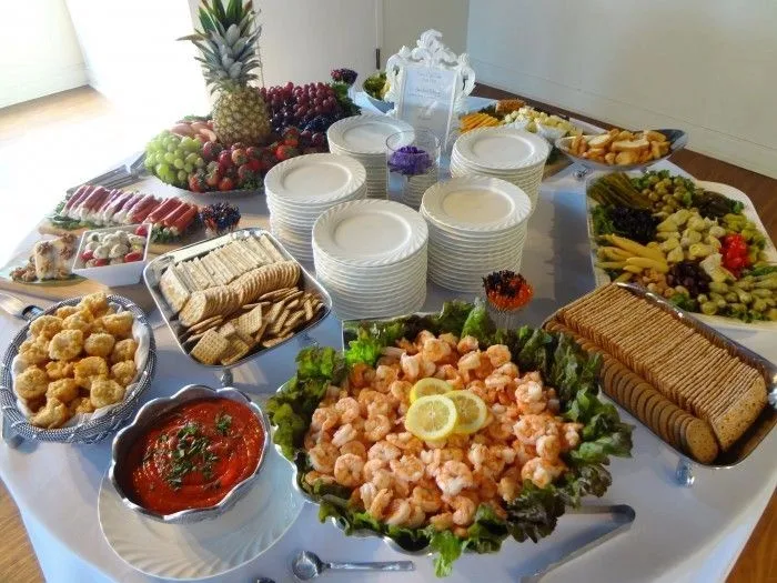 Buffet spread with shrimp on lettuce, crackers, cheese, watermelon, pineapple, grapes, vegetables, and stacked plates.
