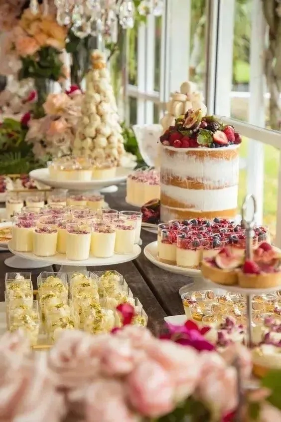 Dessert table featuring a berry-topped naked cake, cream puffs, flower arrangements, and individual cream and berry dessert cups.