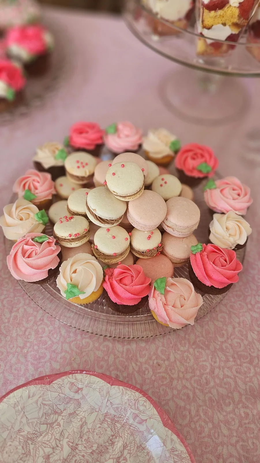 A glass platter displays a pyramid of pale macarons ringed by cupcakes topped with pink, coral.