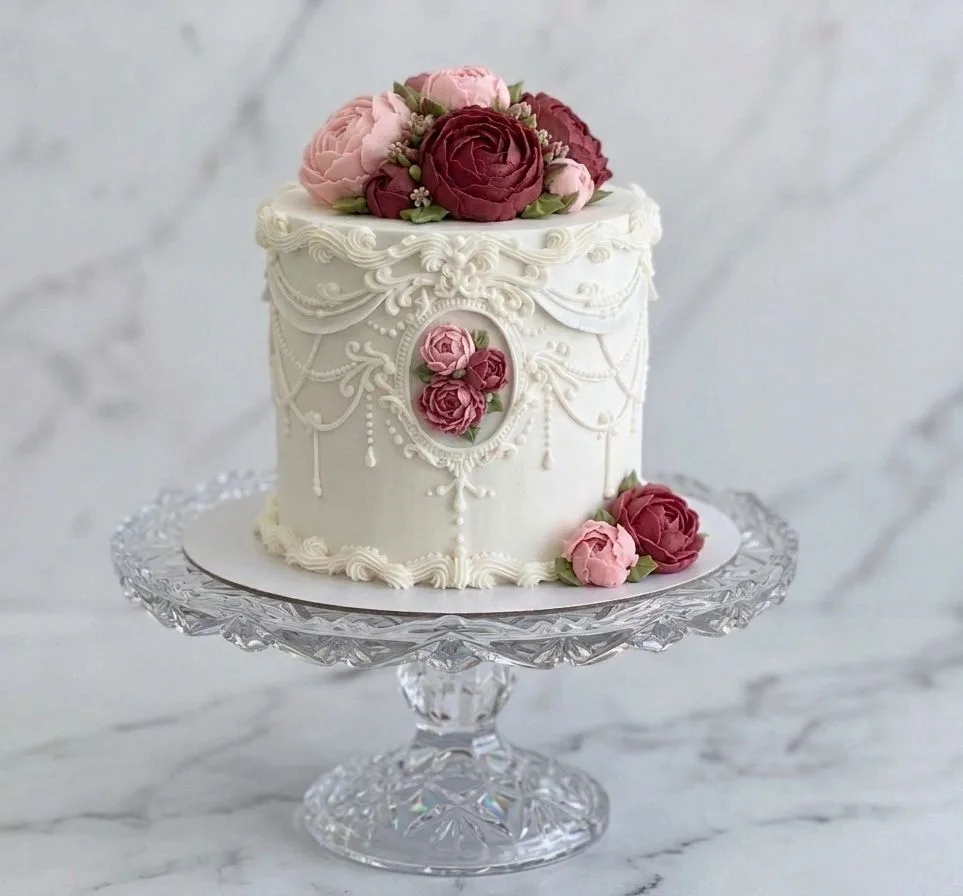Tall white cake with piped lace detailing and pink and burgundy roses displayed on a crystal cake stand.