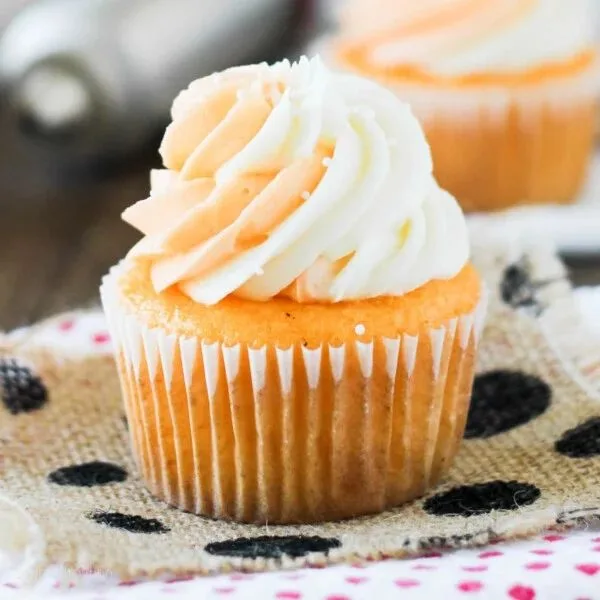 A cupcake with white swirled frosting in a pleated liner rests on burlap with black polka dots. A second, blurry cupcake is visible behind it.