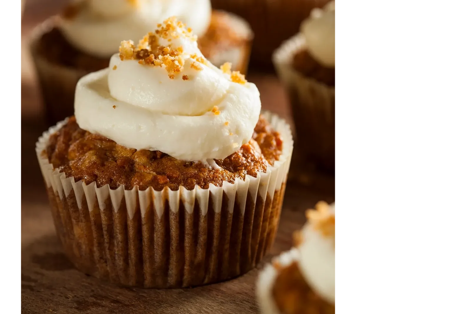 A muffin topped with swirled white frosting and golden crumb sprinkles, sitting in a white paper liner on a wooden surface.