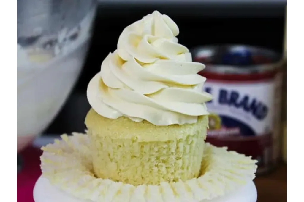 A pale yellow cupcake topped with a tall swirl of white frosting sits on a scalloped white paper liner.