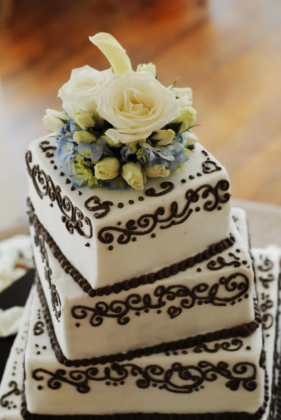 Three-tier square white wedding cake with dark chocolate scrollwork piping, topped with cream roses, blue hydrangea and a white calla lily.