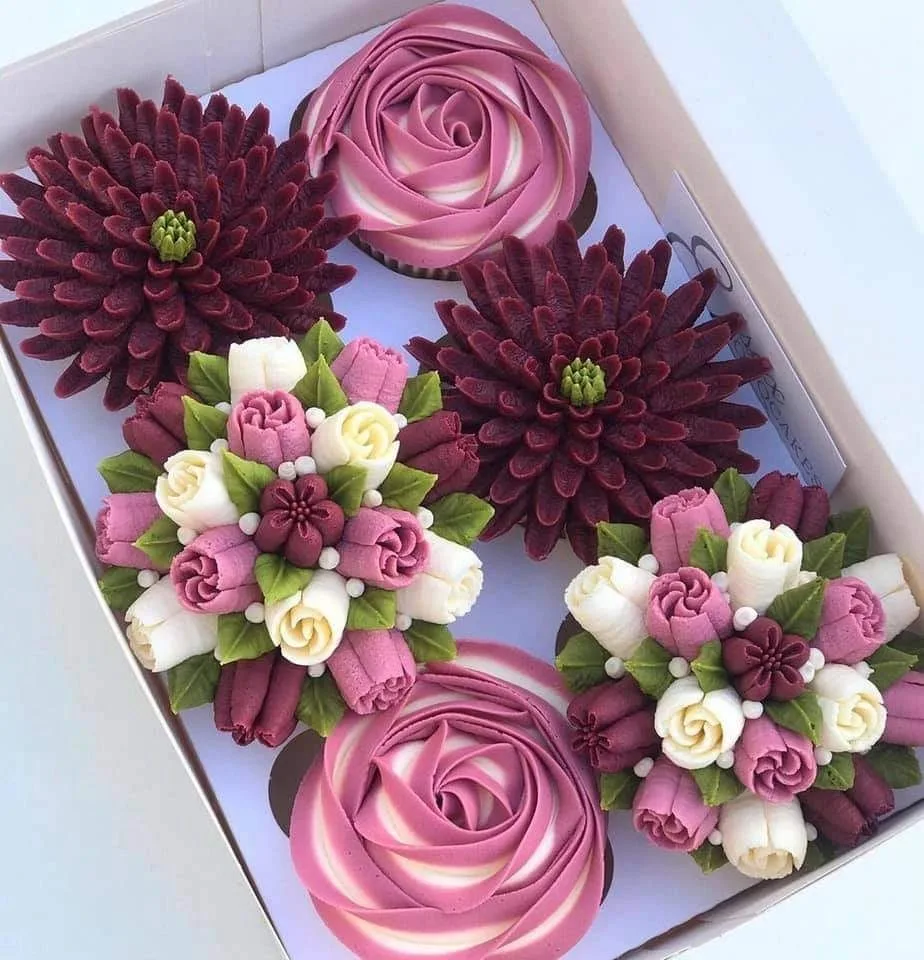 A box of decorated cupcakes featuring pink rose swirls and burgundy dahlia shapes, alongside bouquet designs of piped pink, white.