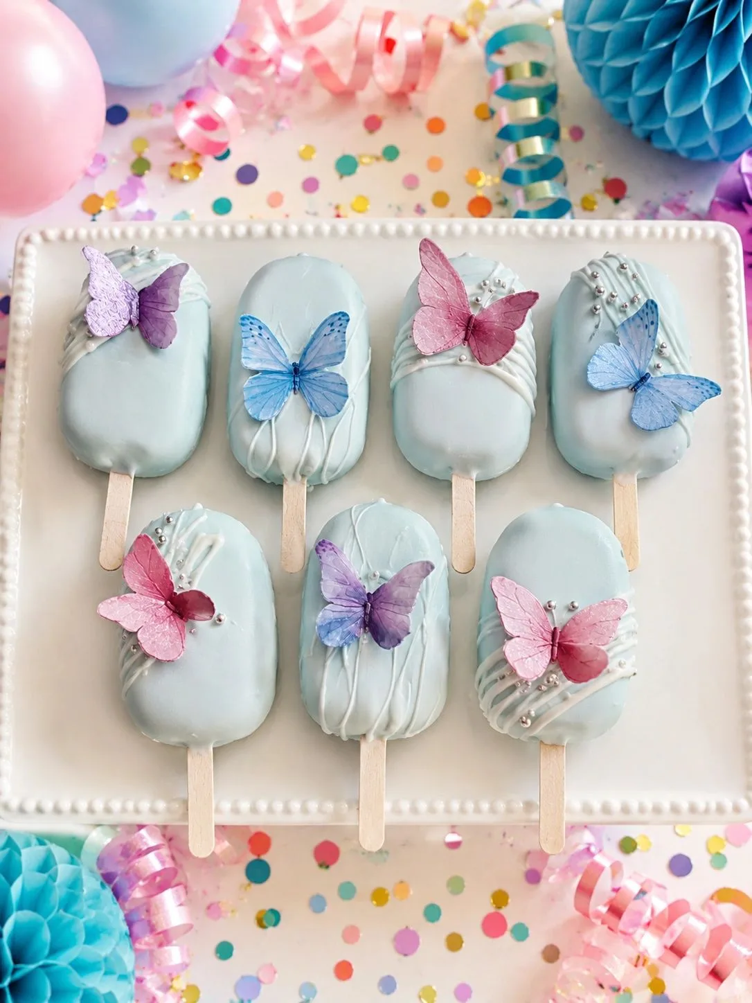 Light blue coated cake pops decorated with pink, purple, and blue edible butterflies on a white tray surrounded by party confetti.
