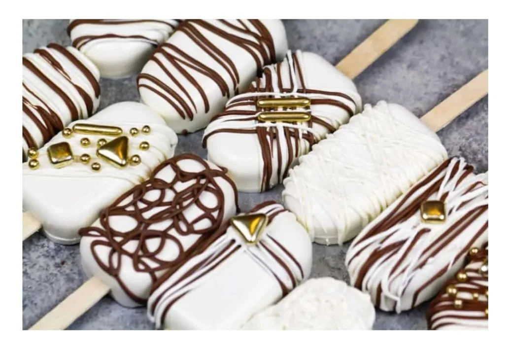 White chocolate-coated treats on sticks decorated with chocolate drizzle patterns and gold geometric accents arranged on a gray surface.