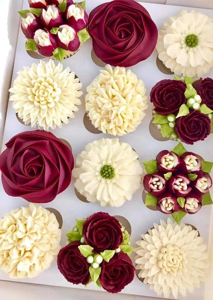 A box of twelve cupcakes decorated with buttercream frosting piped to resemble burgundy roses, white dahlias, and chrysanthemums with green leaves.