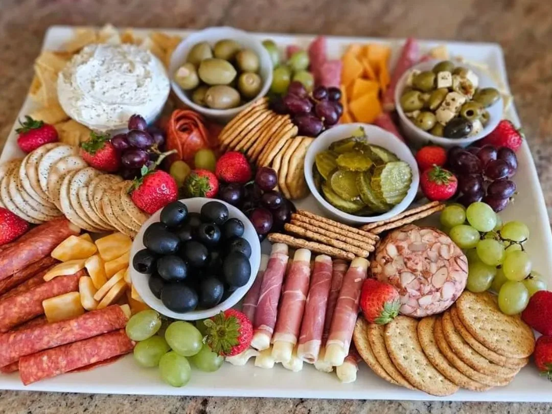 Charcuterie board with crackers, grapes, strawberries, olives, pickles, salami, cheese cubes, pretzels, and a nut-coated cheese ball.