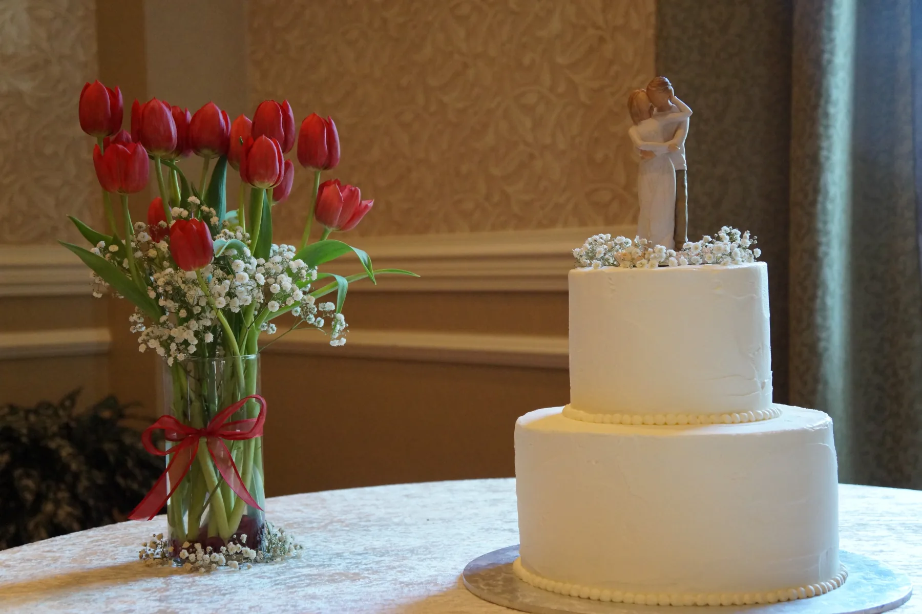 Two-tier white buttercream wedding cake with a bride and groom topper and baby&rsquo;s breath, beside a glass vase of red tulips.
