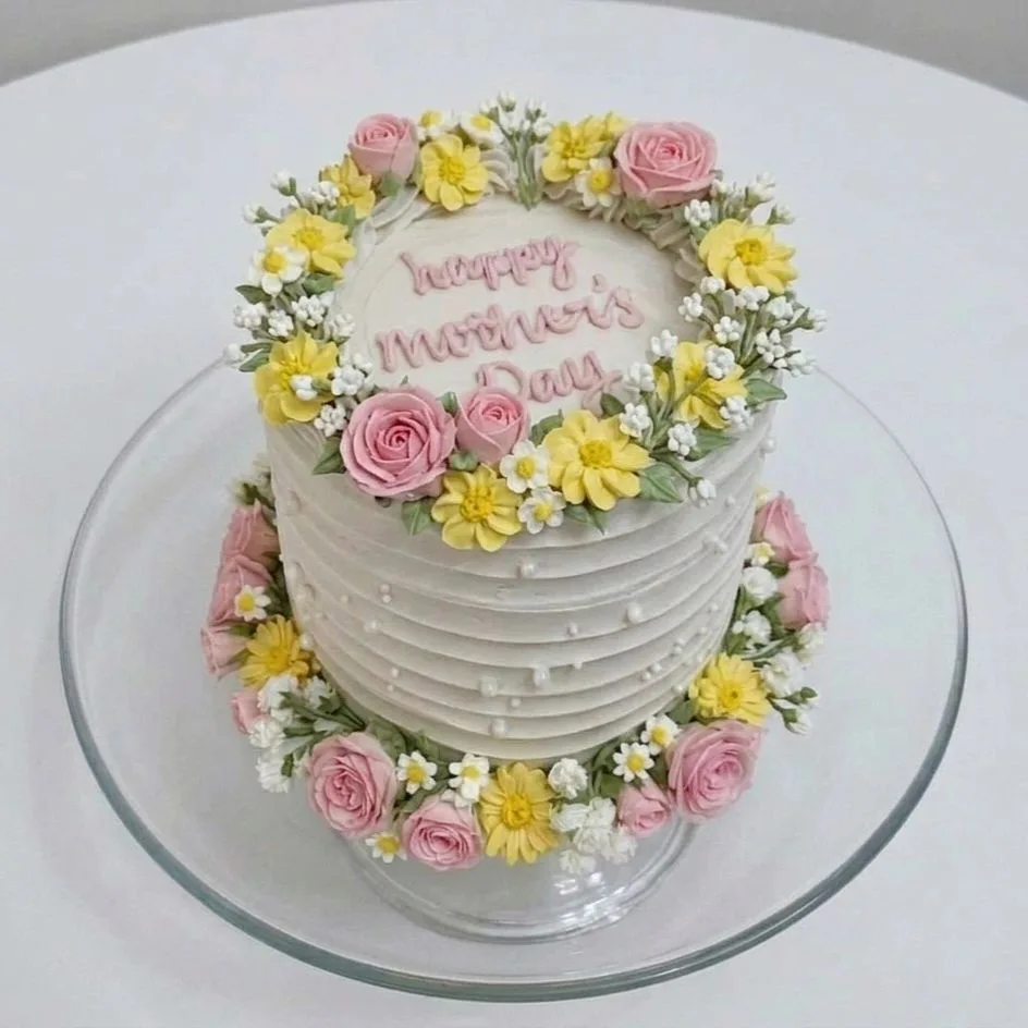 White frosted round cake on a glass stand, decorated with pink and yellow buttercream flowers and 
