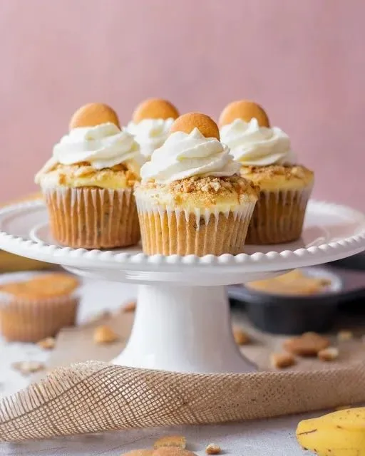Four cupcakes topped with swirled white frosting and round vanilla wafer cookies sit on a white pedestal cake stand.