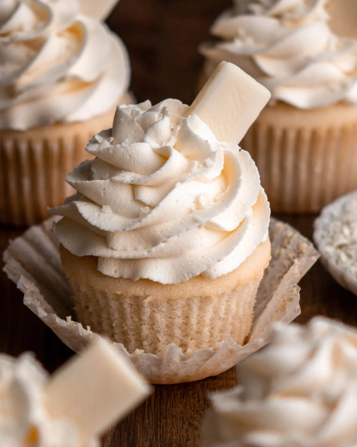 Vanilla cupcakes in brown paper liners topped with swirled white buttercream frosting and a white chocolate stick, arranged on a wooden surface.